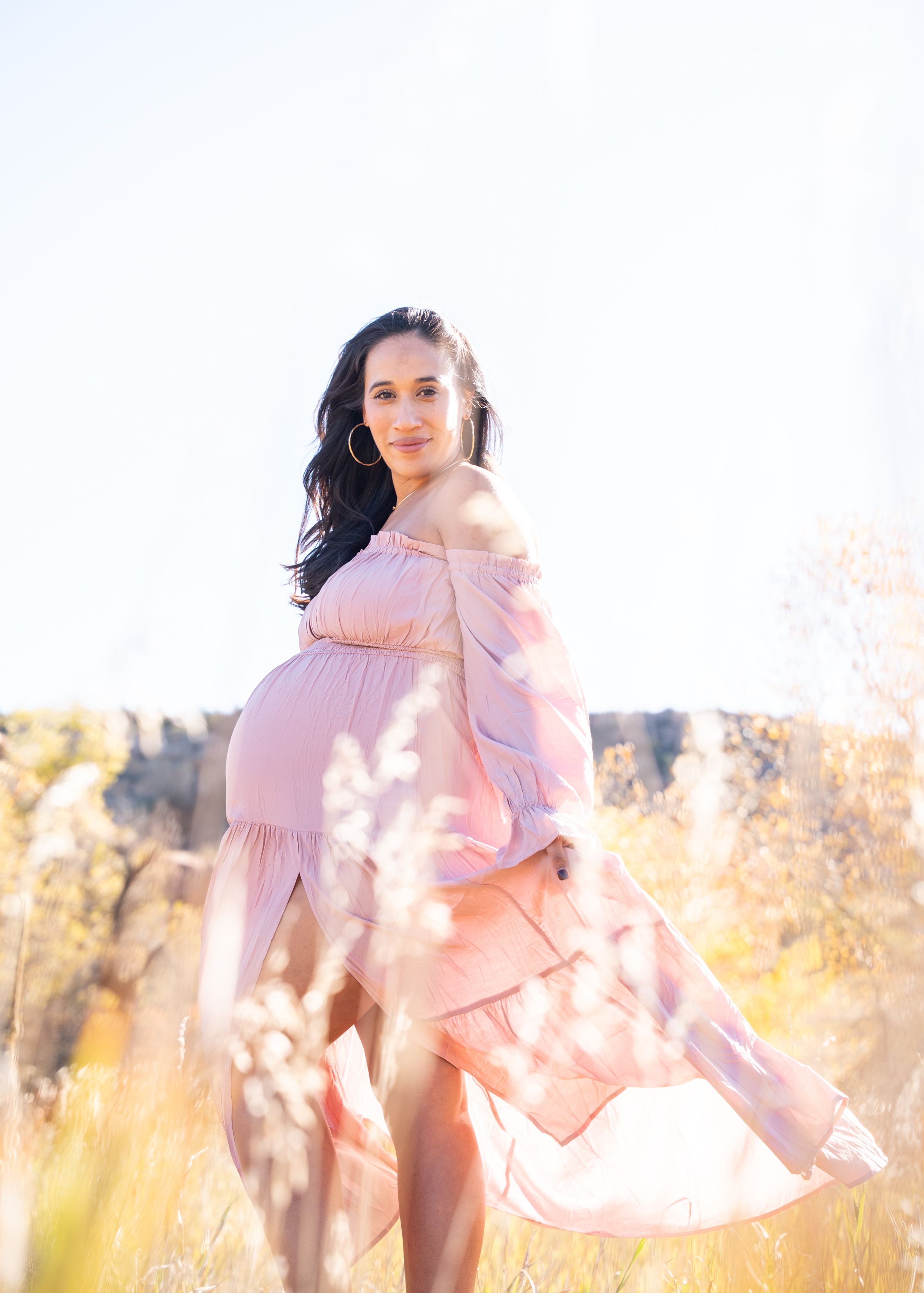 Woman in flowing pink dress poses gracefully in golden sunlit field with tall grass and dreamy atmosphere.