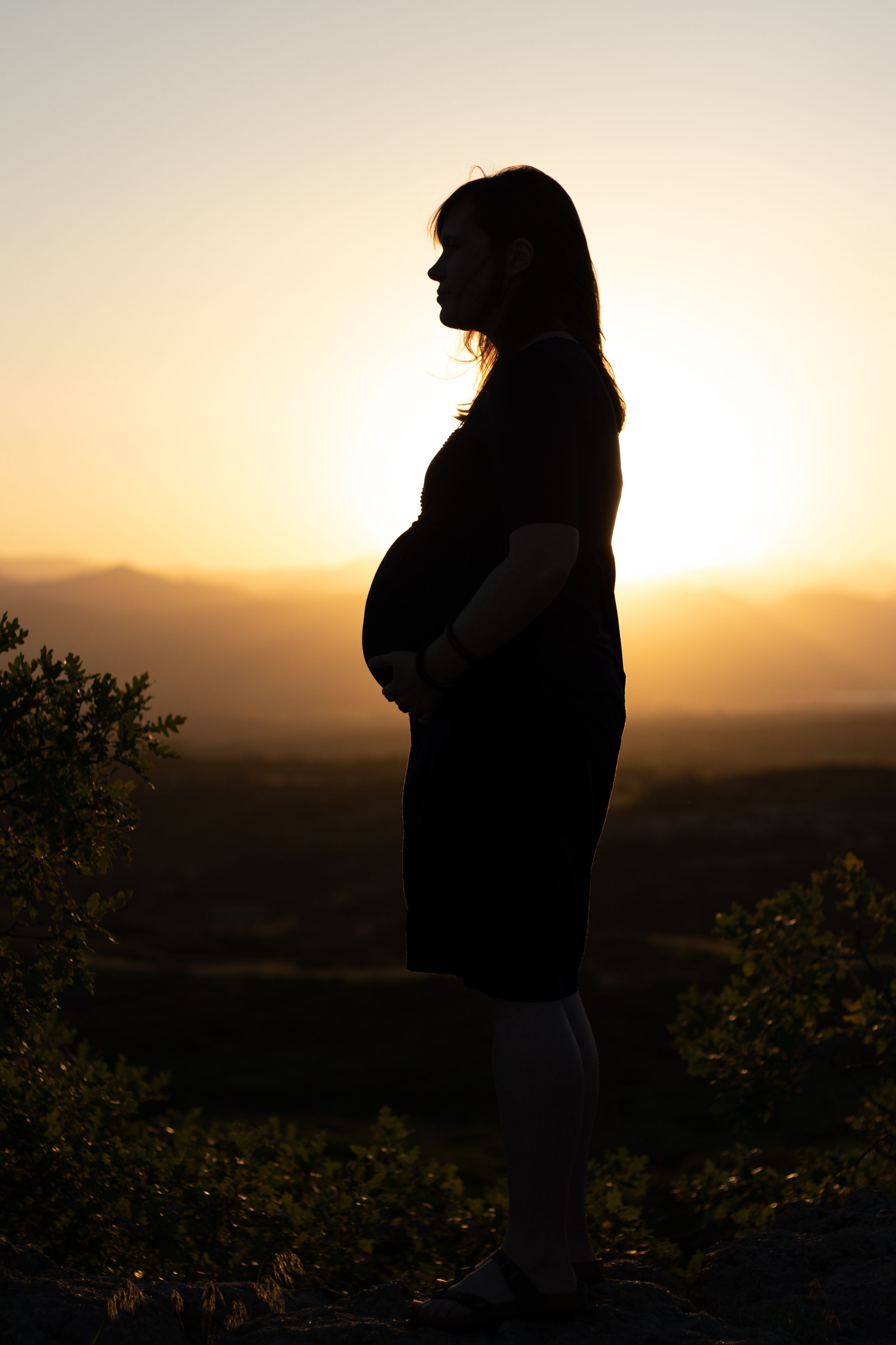 Side profile silhouette of pregnant mother at sunset with mountains in background.