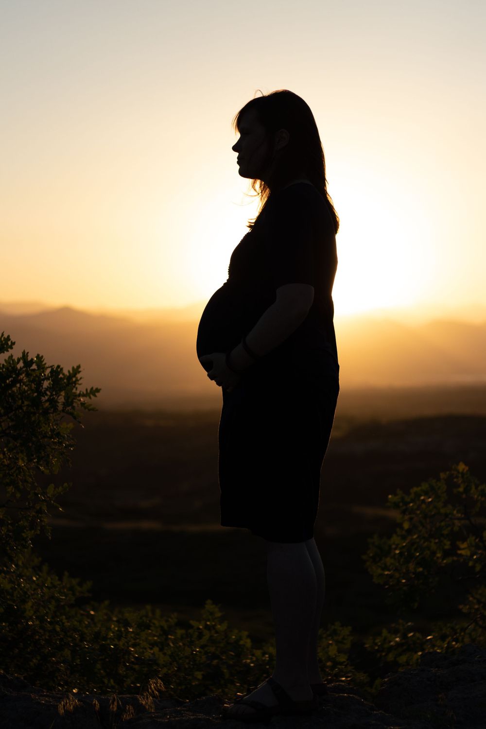Side profile silhouette of pregnant mother at sunset with mountains in background.