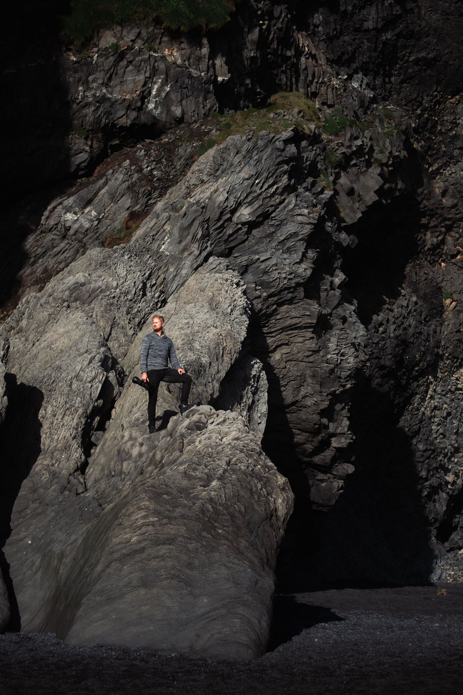 Person standing on dramatic rocky cliff formation near dark cave entrance.