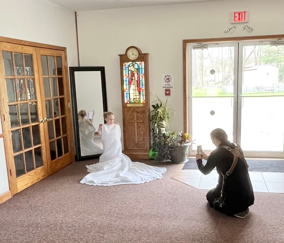 Young girl in white dress poses near mirror while photographer kneels to capture photo in church foyer.