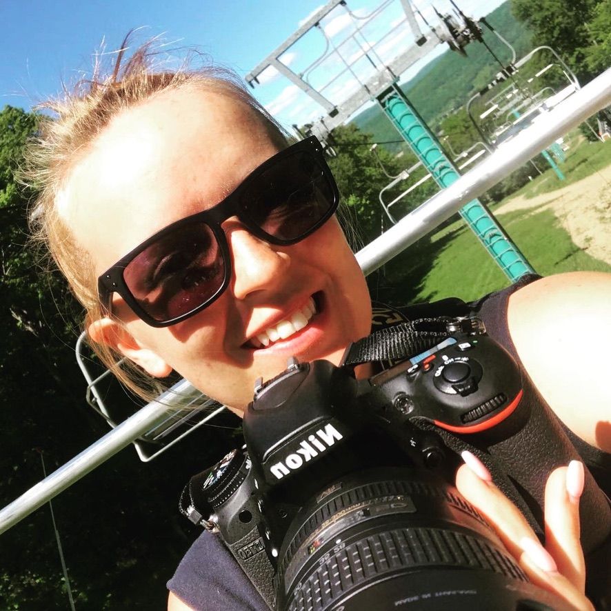 Smiling woman taking a selfie outdoors while holding a Nikon camera, wearing sunglasses near a ski lift on a sunny day.