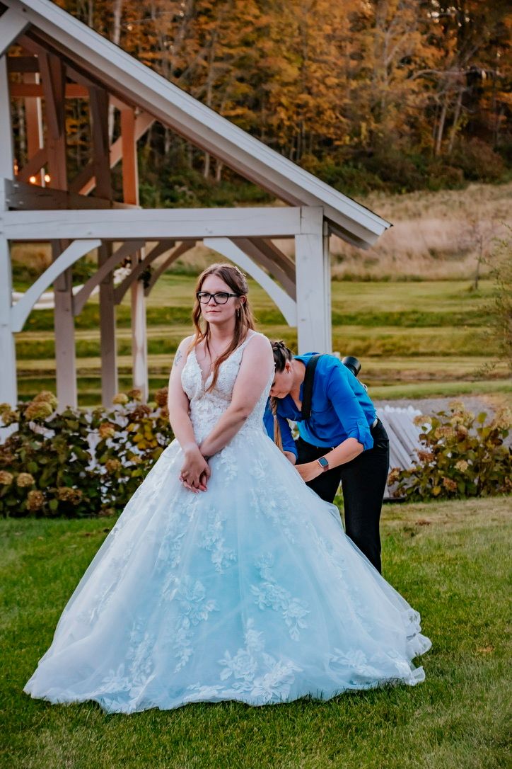 Bride in white lace ball gown being helped by assistant outdoors near wooden arbor with autumn trees in background.