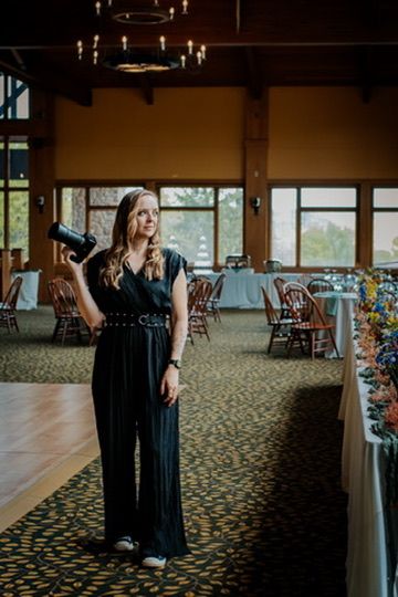 A female photographer in a black jumpsuit holds a camera while standing in an elegant event venue with chandeliers.