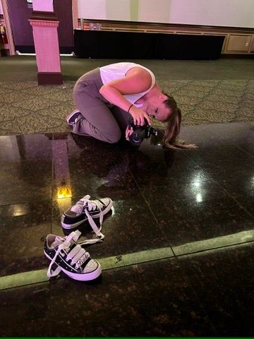 Woman crouching on shiny floor photographing sneakers in dimly lit venue with patterned carpet.