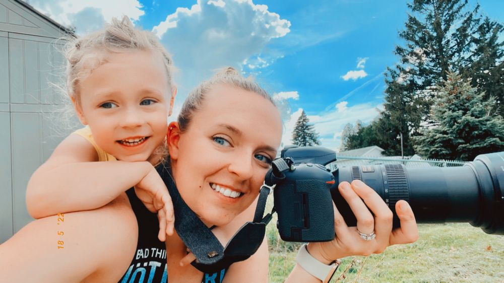 Woman smiling holding a camera outdoors with a young blonde child on her shoulders under a blue sky.