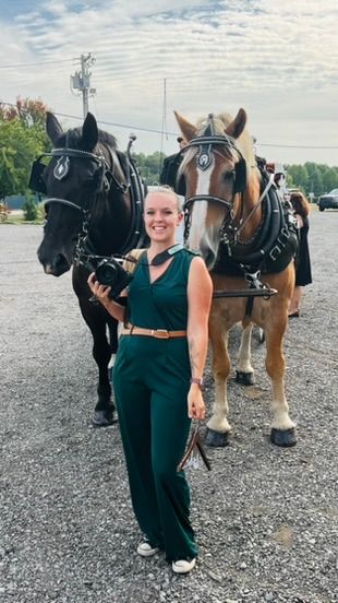 Woman in green jumpsuit smiling beside two harnessed draft horses on a gravel lot.