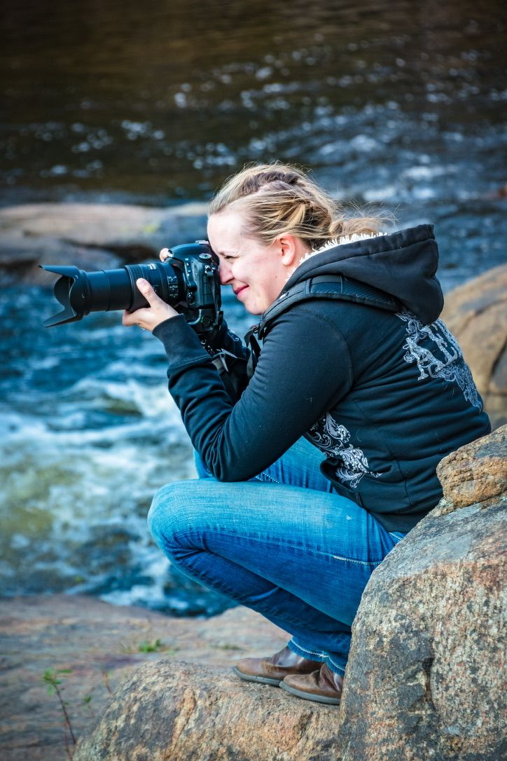 Woman crouching on rocks by a river, photographing with a DSLR camera and telephoto lens, wearing a black hoodie.