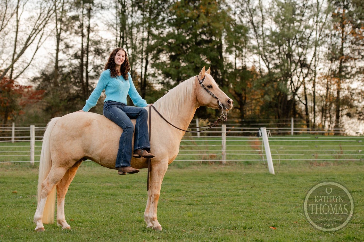 A rider sits bareback on a cream-colored horse in a fenced pasture during autumn.