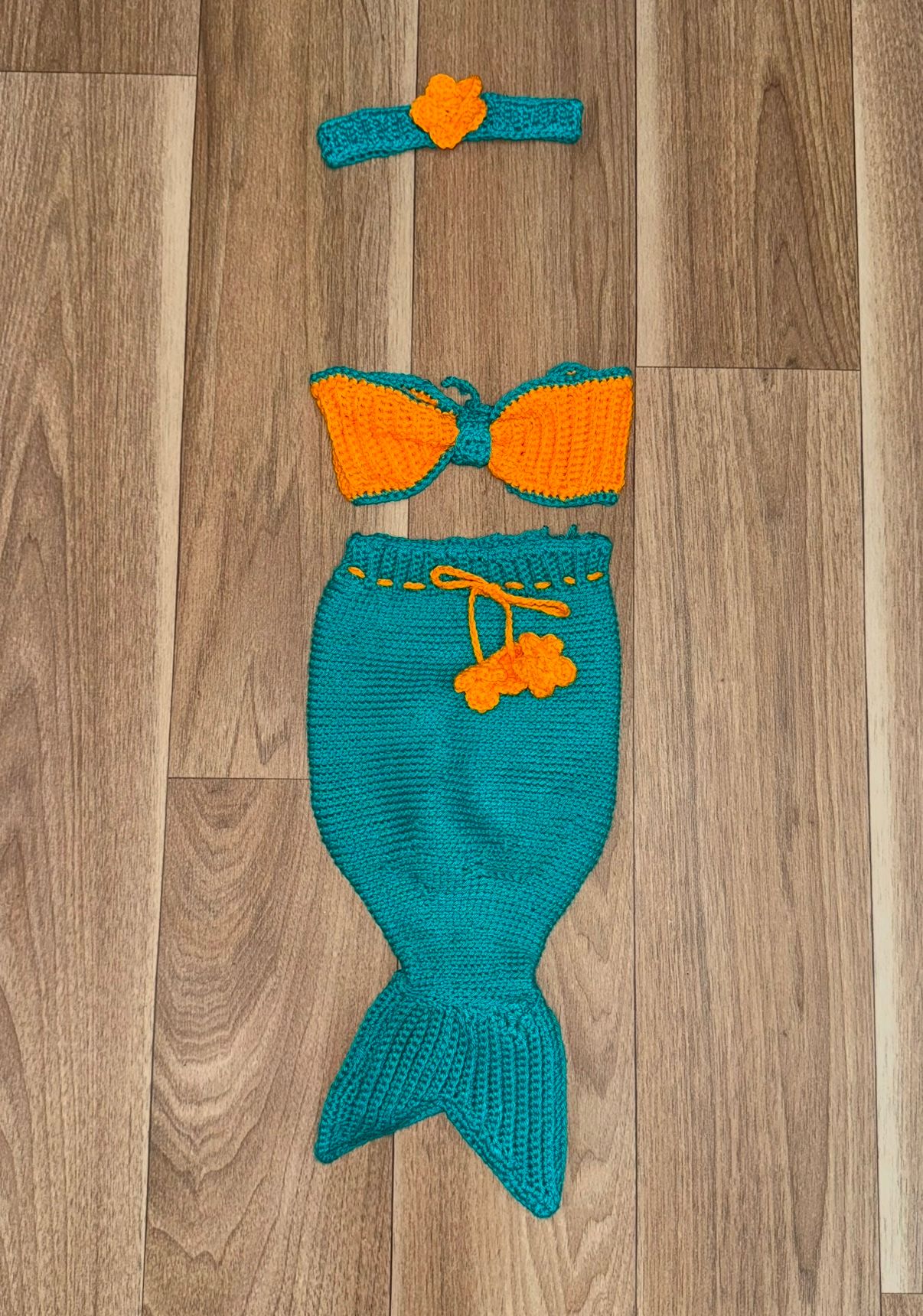 Teal and orange crochet mermaid tail outfits displayed on wooden floor.