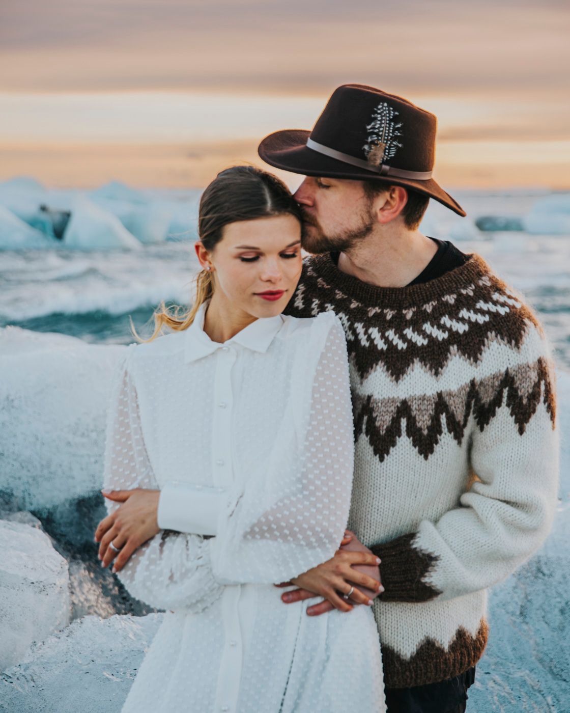 Romantic couple embraces near glacial ice formations wearing nordic sweater and white dress at sunset.