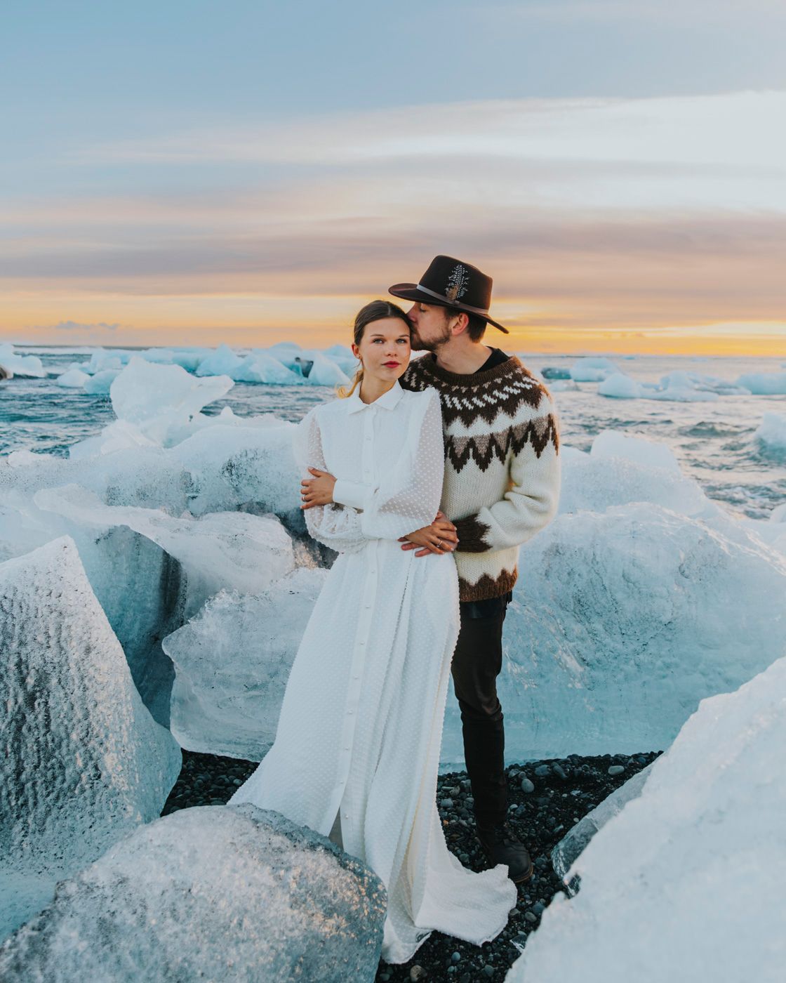 A couple embraces among blue glacial ice formations at sunset, wearing winter attire against an orange and pink sky.