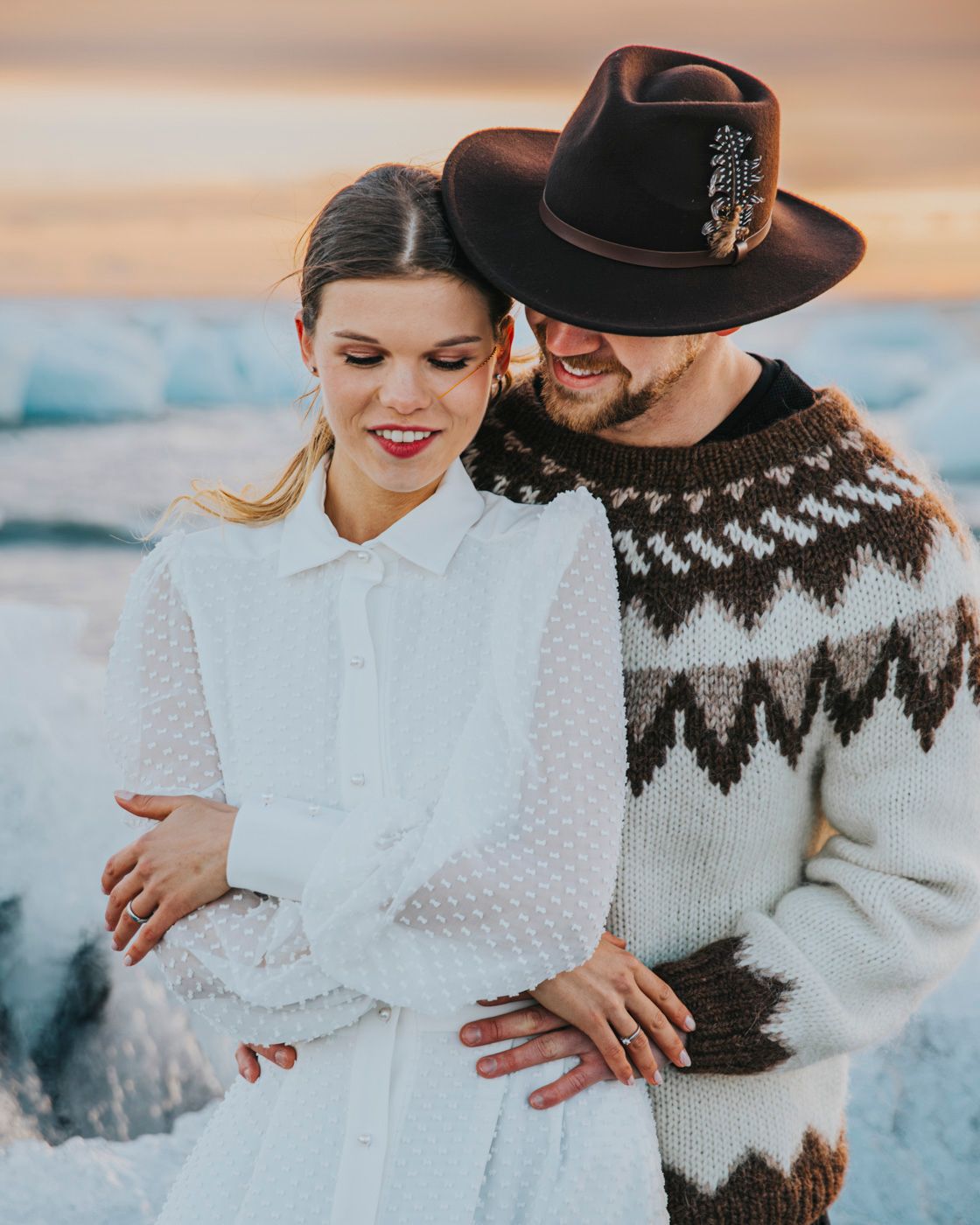 Couple embracing in winter attire with person wearing nordic sweater and brown fedora against icy blue background.