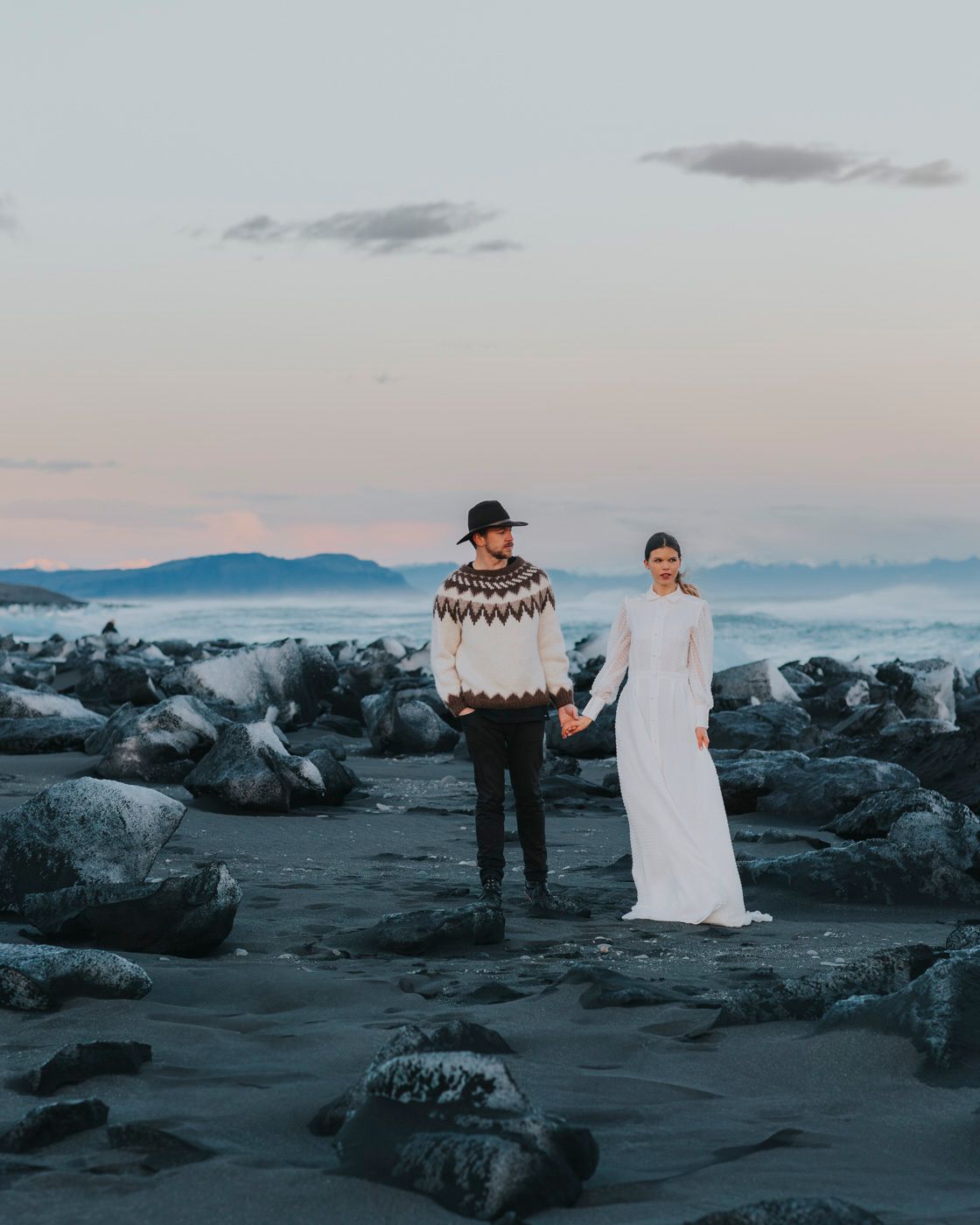 Couple in nordic attire poses together on rocky beach with misty mountains in background at dusk.