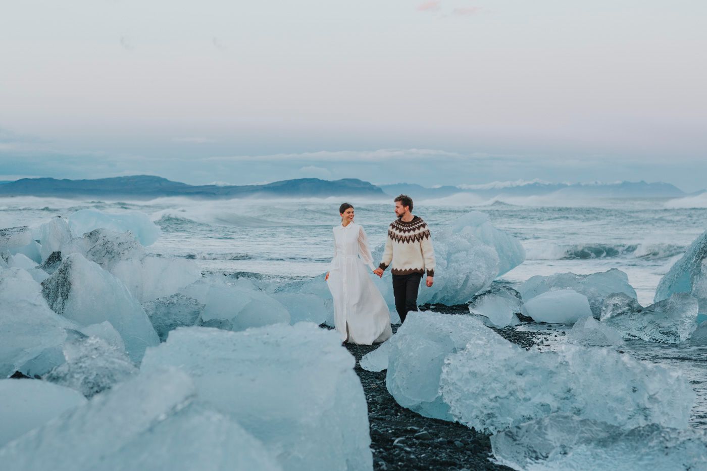 Two people stand on a stunning glacial beach with towering ice formations in Iceland during sunset.