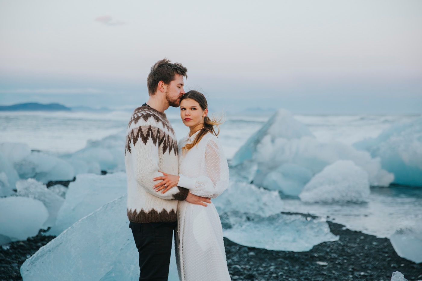 Two people share a romantic moment on a beach with dramatic ice formations and blue glacial ice in the background.