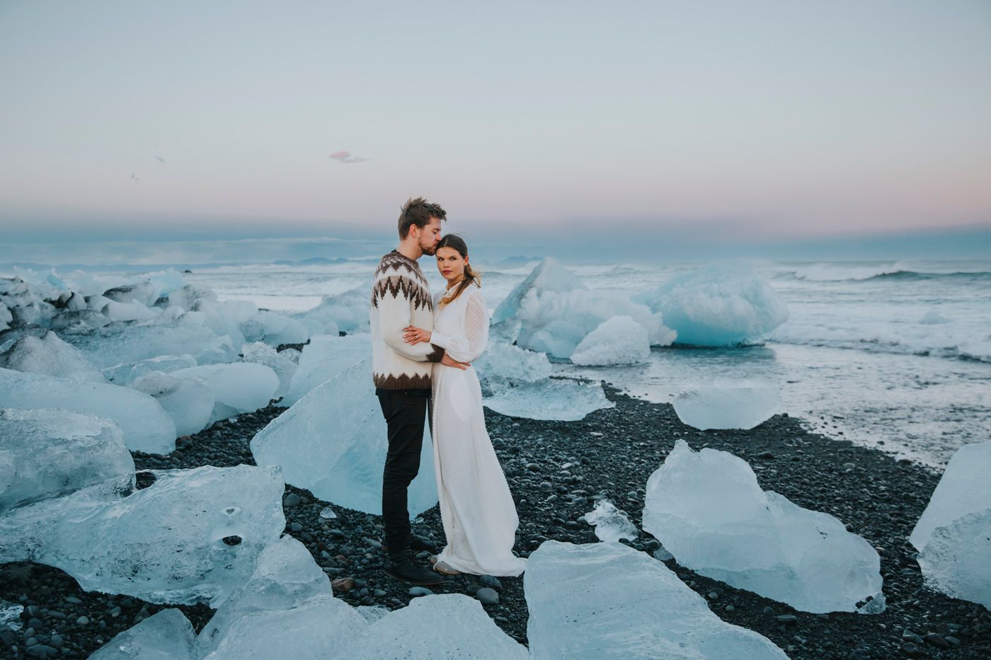 A couple in wedding attire embrace on a black sand beach surrounded by dramatic blue ice formations at dusk.