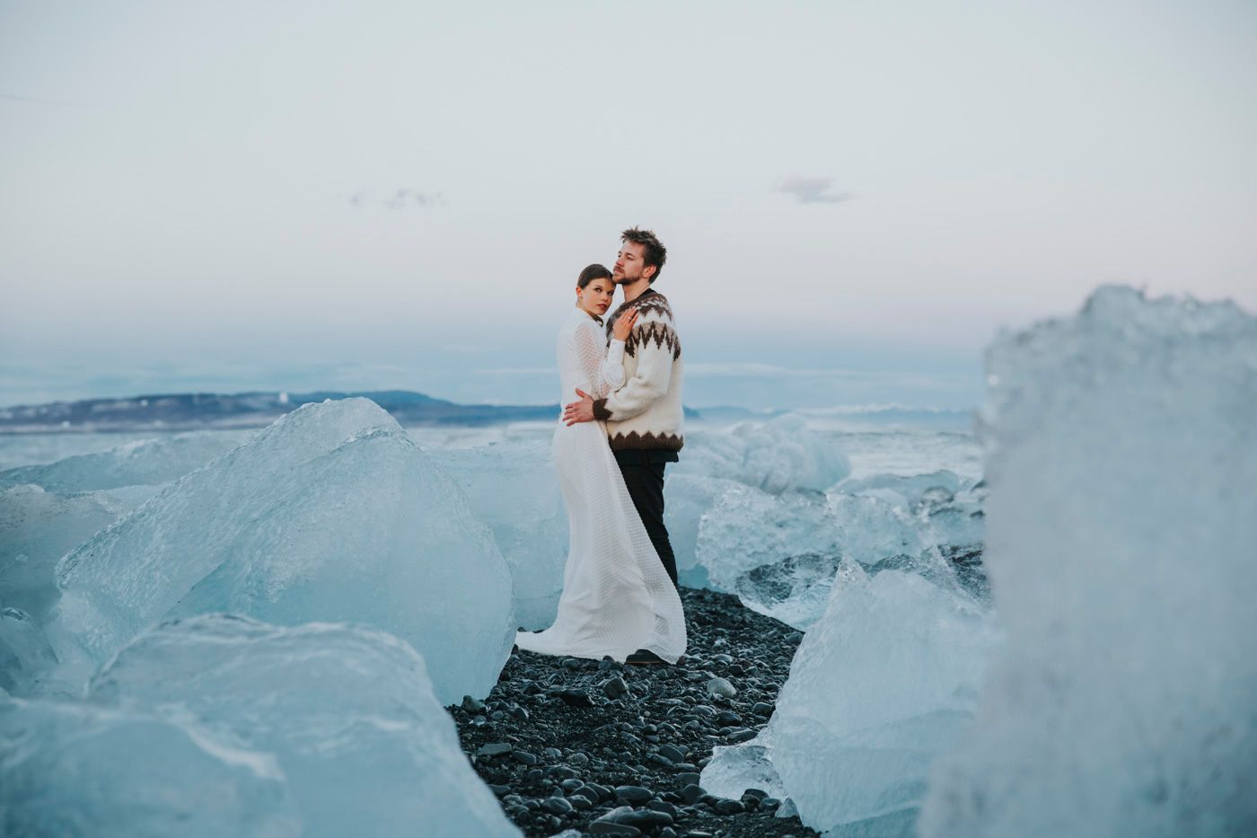 A couple in wedding attire embrace while standing on black sand beach surrounded by dramatic blue ice formations in Iceland.
