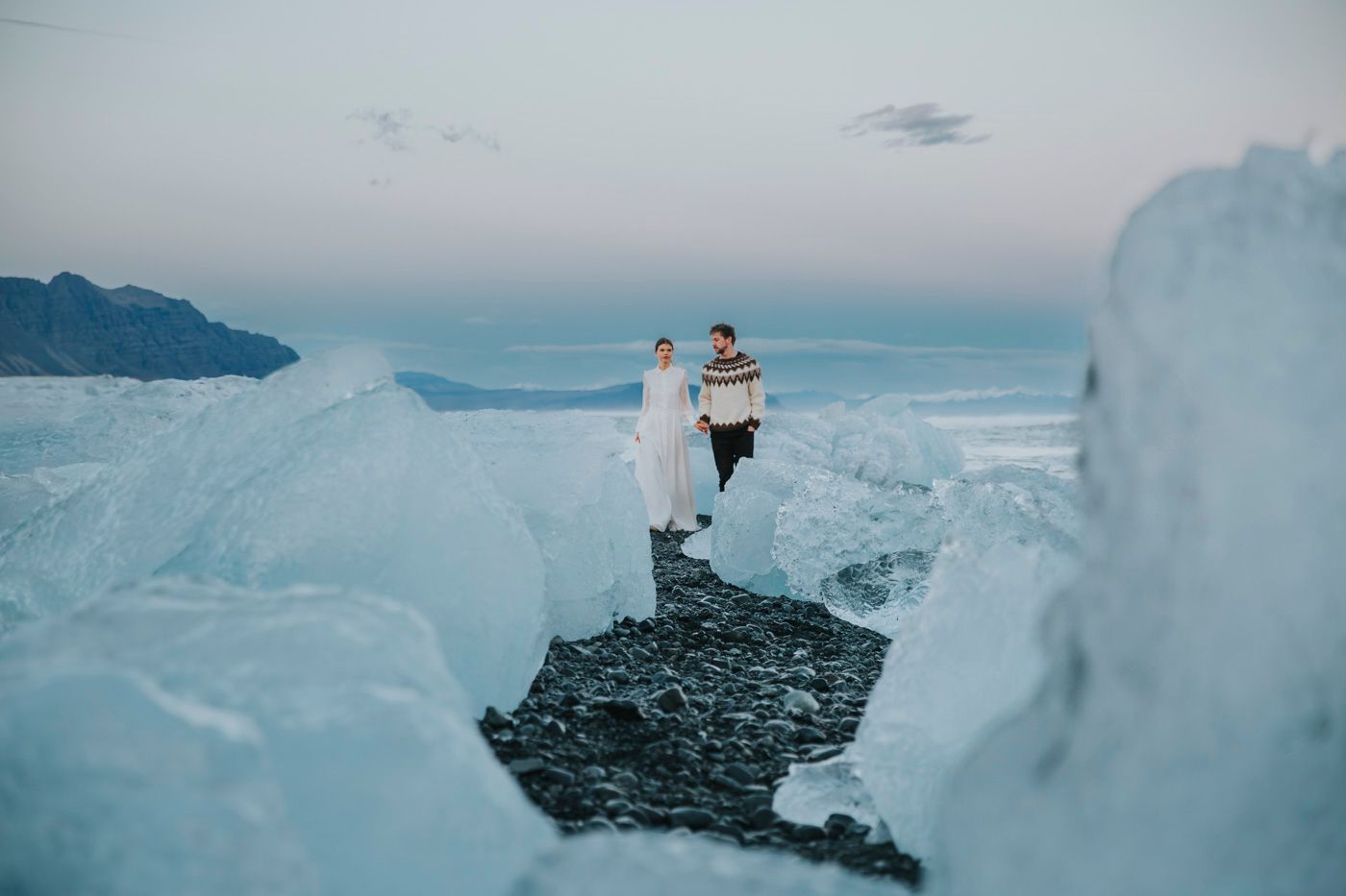 A couple stands atop a black rock surrounded by dramatic ice formations at Diamond Beach in Iceland during sunset.