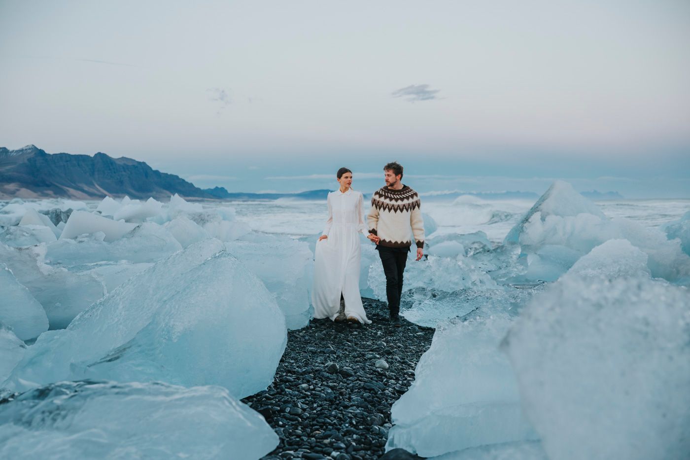 Two people in winter attire stand on dark rocks surrounded by dramatic blue glacier ice formations in Iceland.