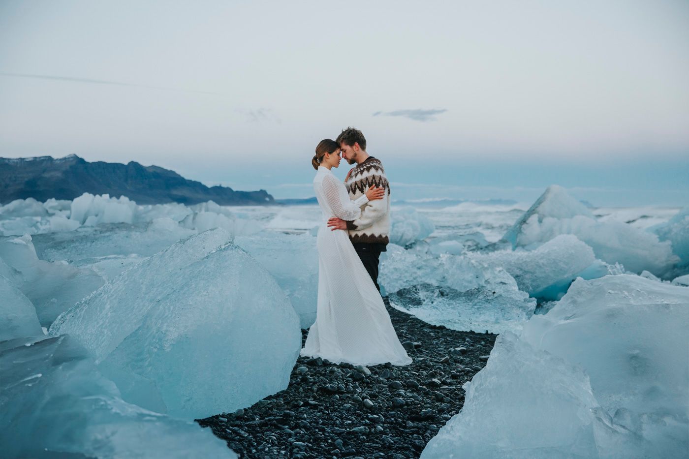 Two people embrace on a rocky beach surrounded by dramatic blue glacier ice formations at dusk.