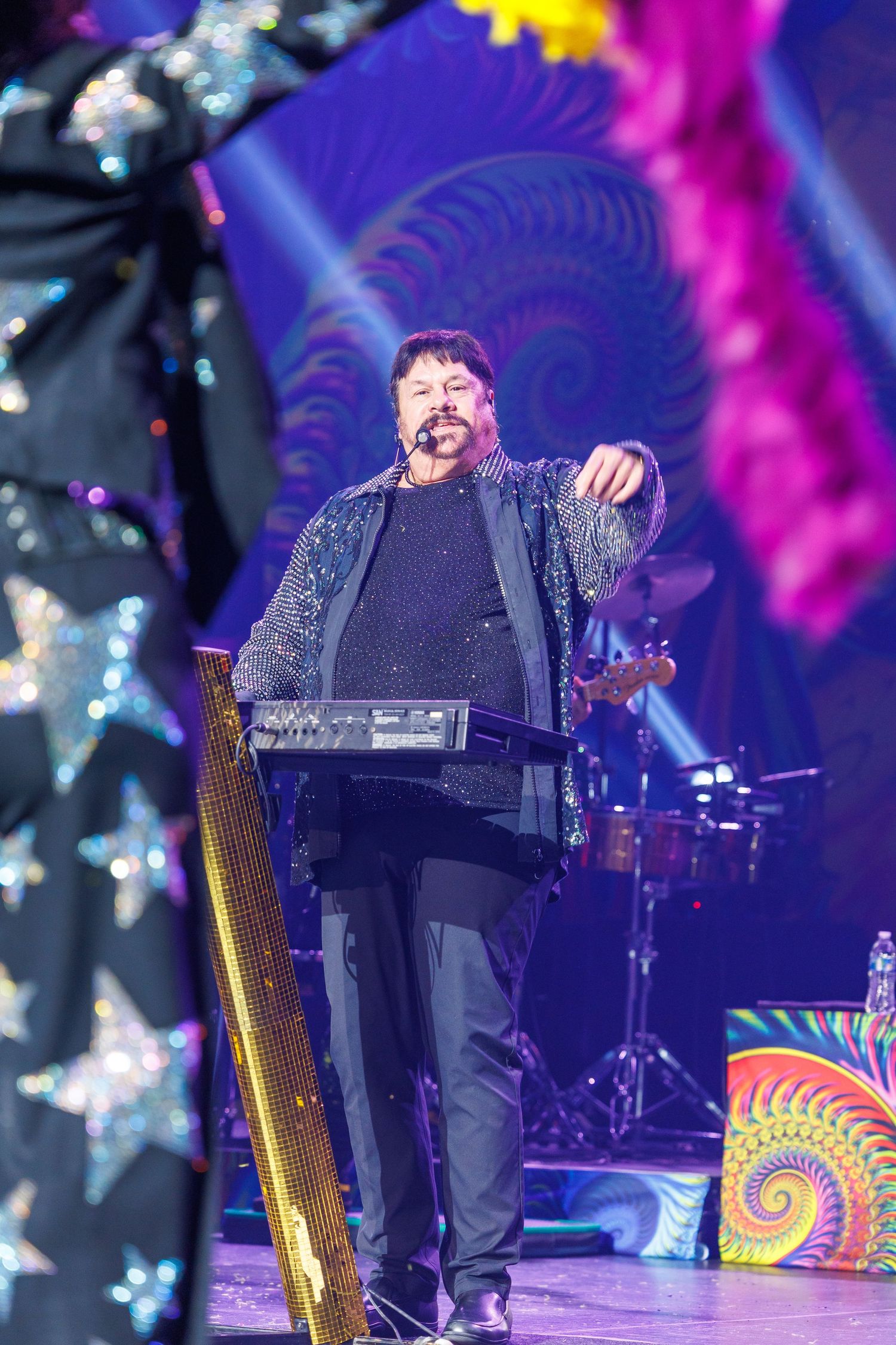 Musician performs on stage with colorful psychedelic backdrop and harp visible during an energetic concert performance.