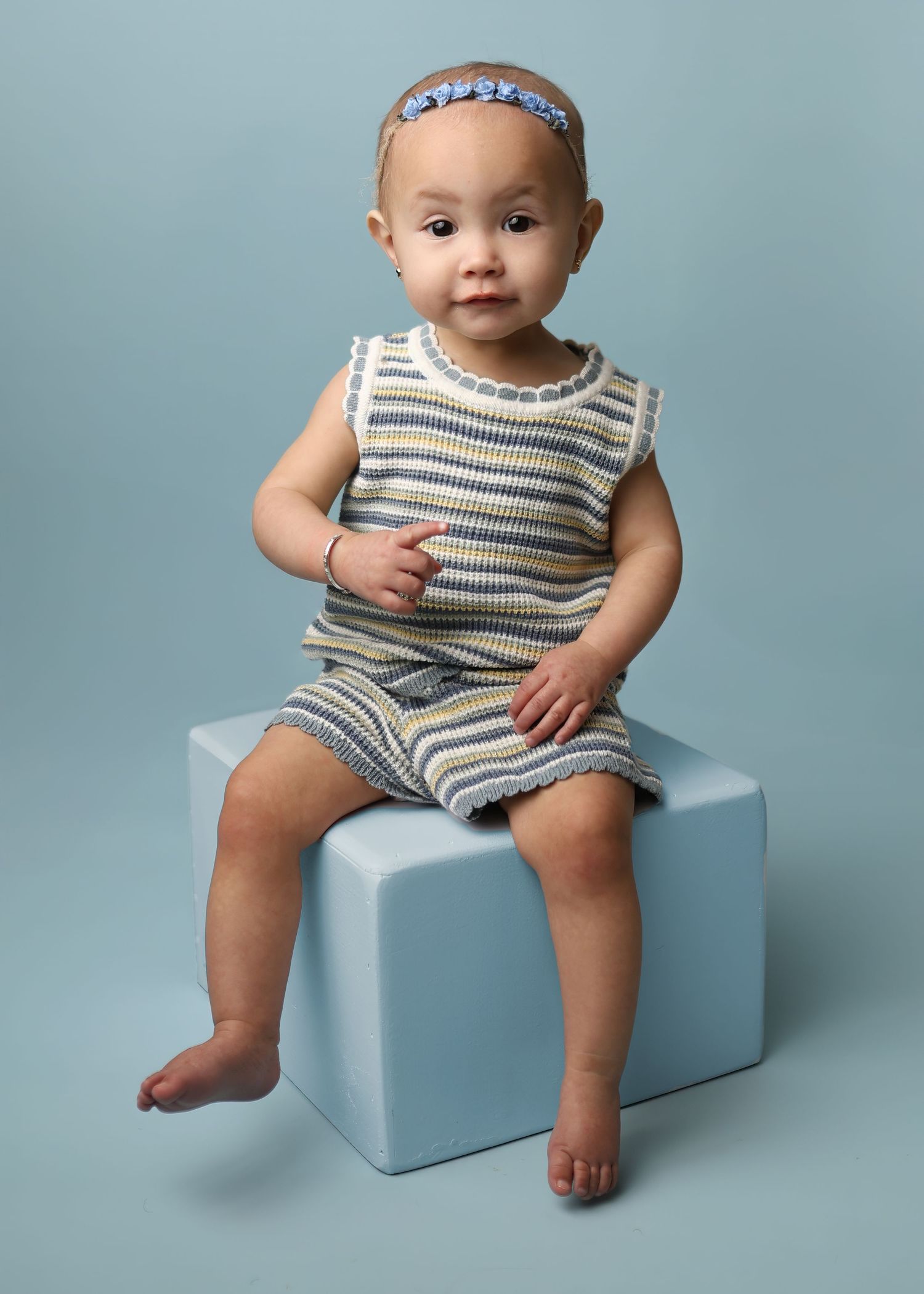 Small child in striped onesie and headband sitting on light blue cube against blue backdrop.