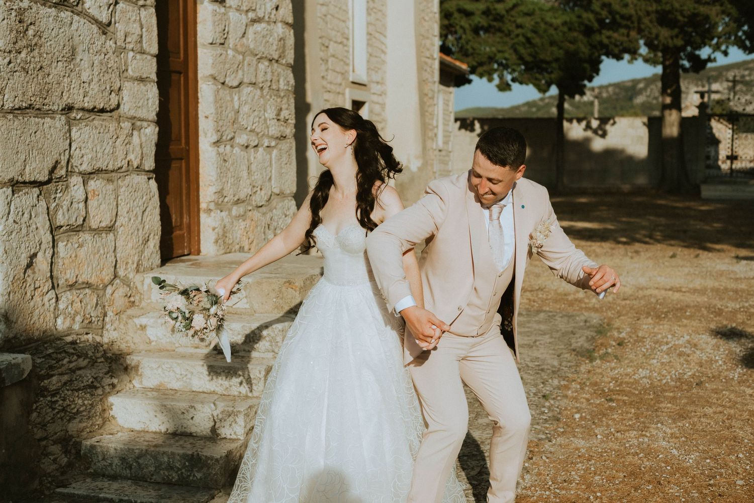 Wedding couple running joyfully outside stone church in sunlight.