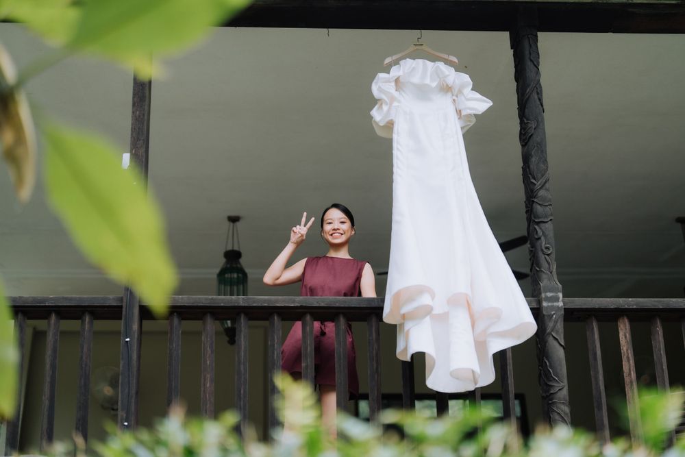 A white wedding dress hangs on display on a porch railing while someone stands nearby in burgundy.
