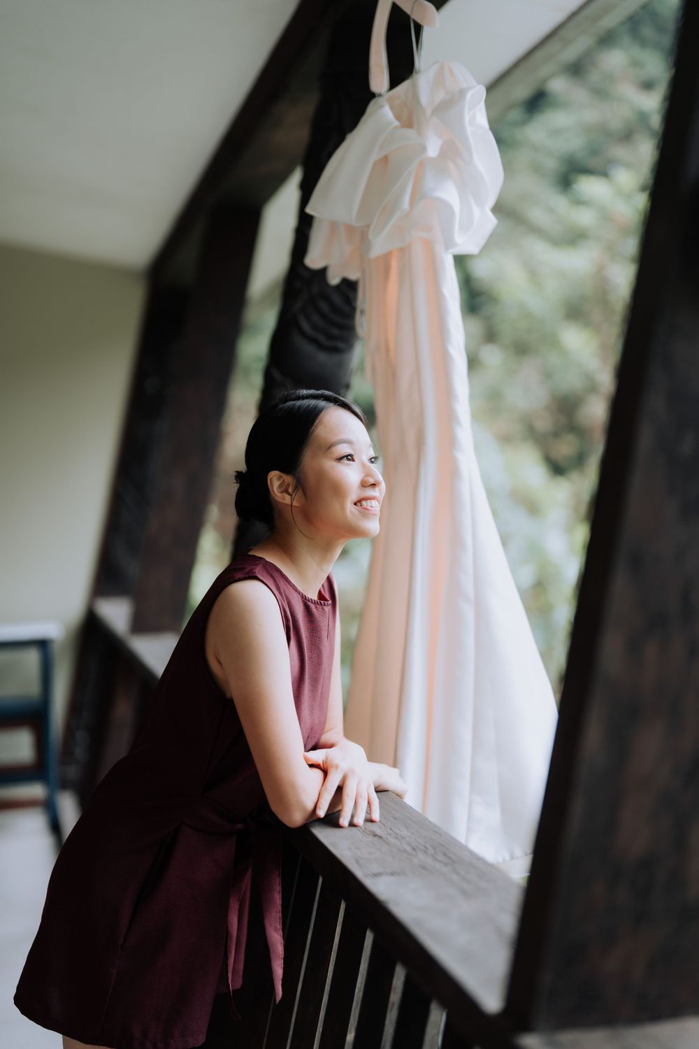 A person in a burgundy dress leans on balcony railing while looking out window with wedding gown hanging above.
