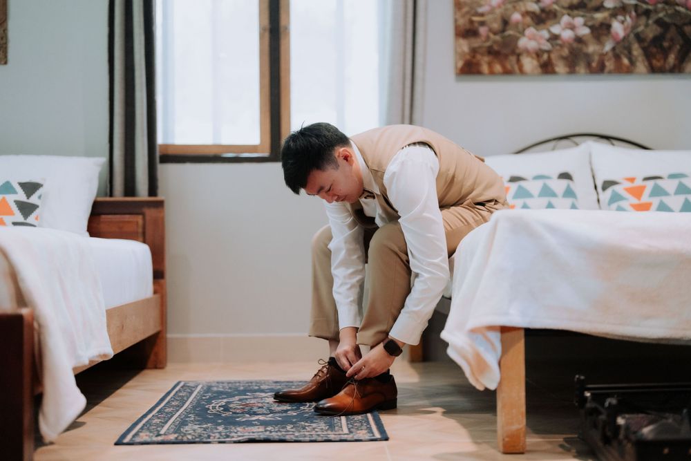 Someone wearing a beige suit jacket leans down to tie their brown dress shoes in a light-filled room with white beds.