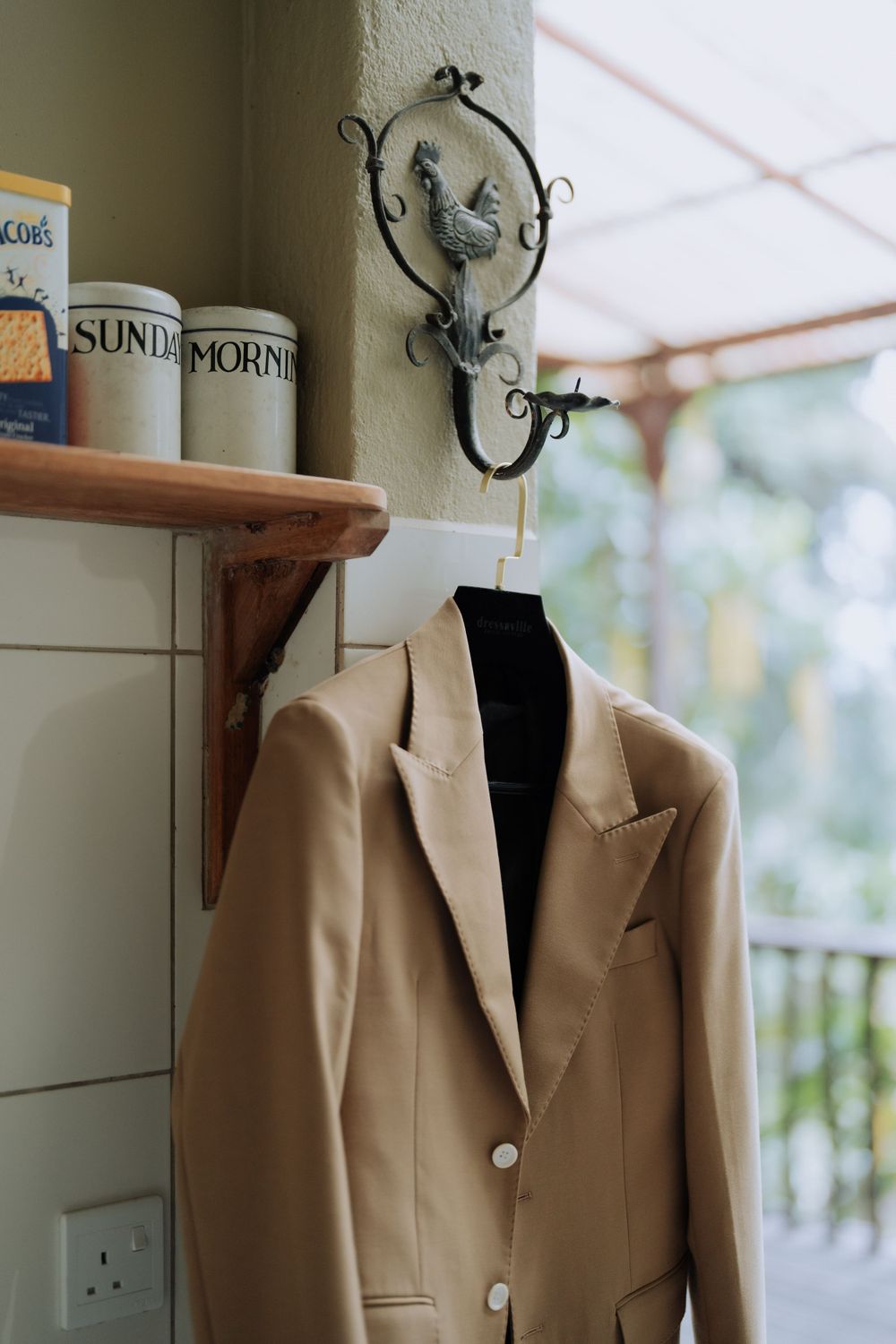 A beige blazer jacket hanging on an ornate wall hook next to a wooden shelf in a home interior.