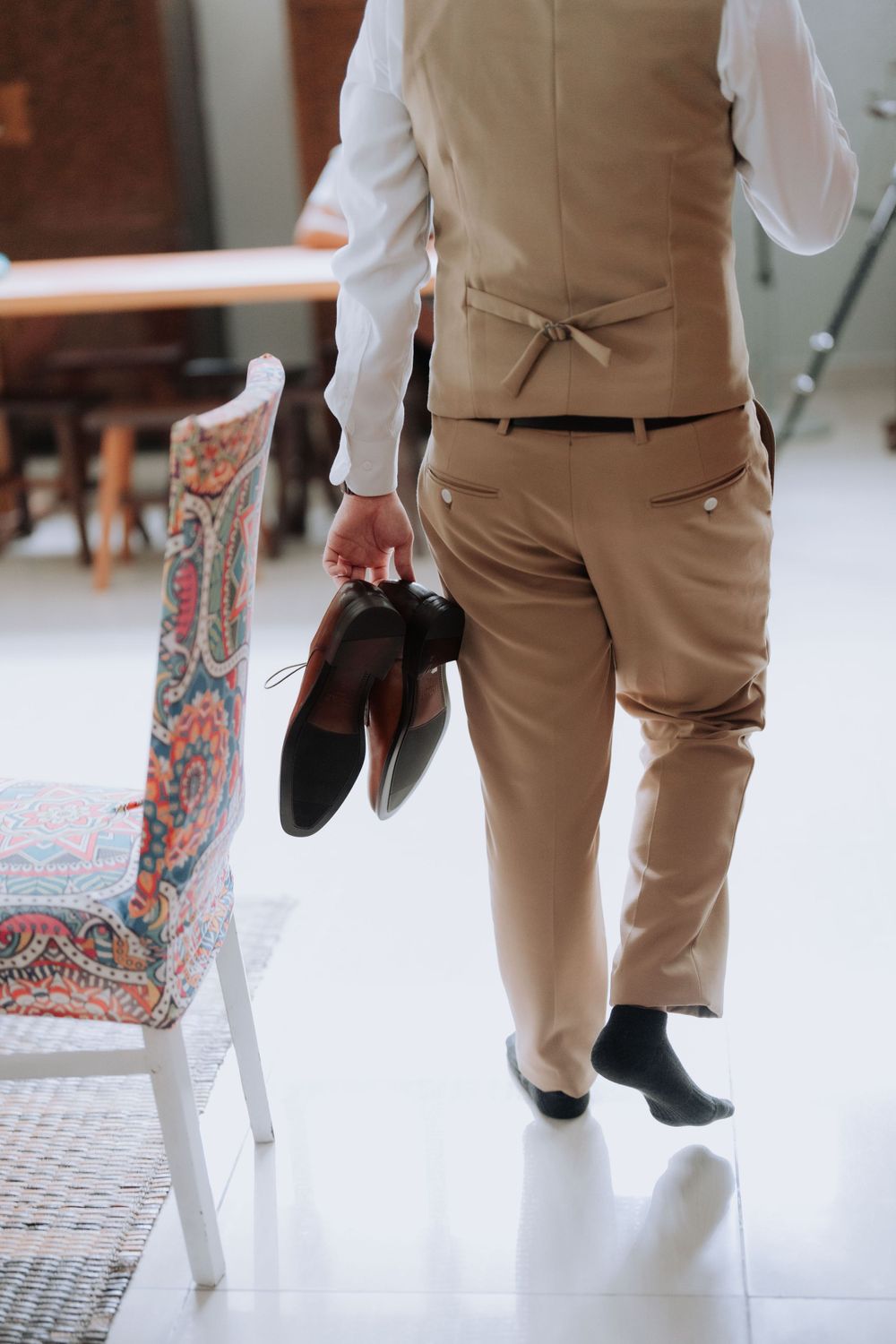 Groom in beige suit holding dress shoes while standing next to a decorative chair at a wedding venue.