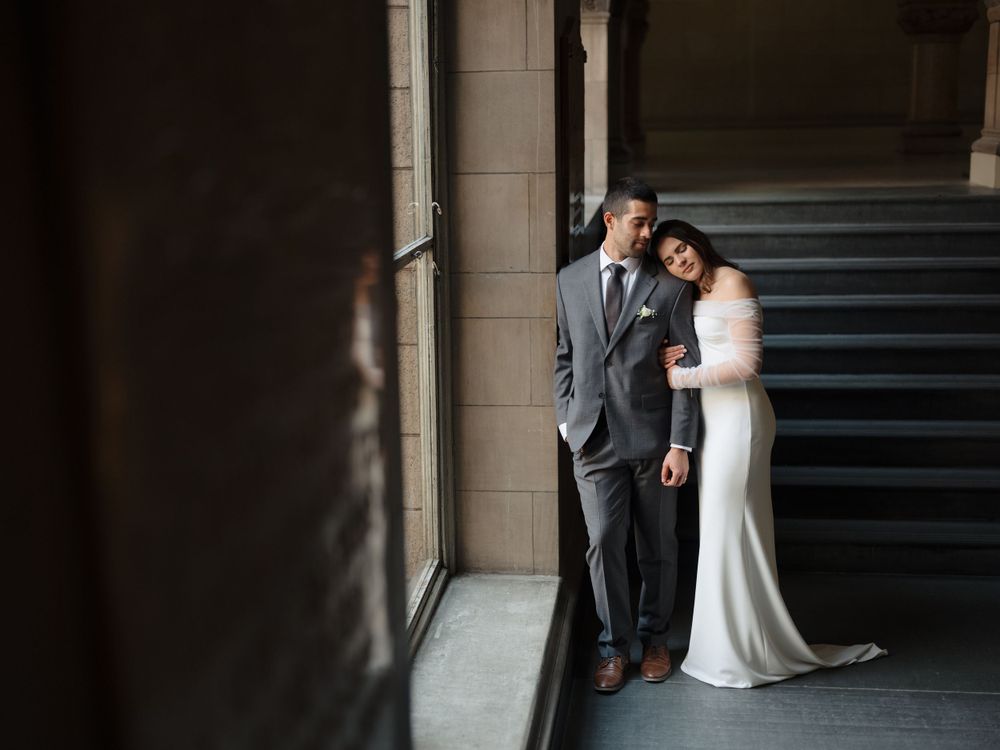 A couple in formal wedding attire stands together in an elegant columned hallway with dramatic lighting.