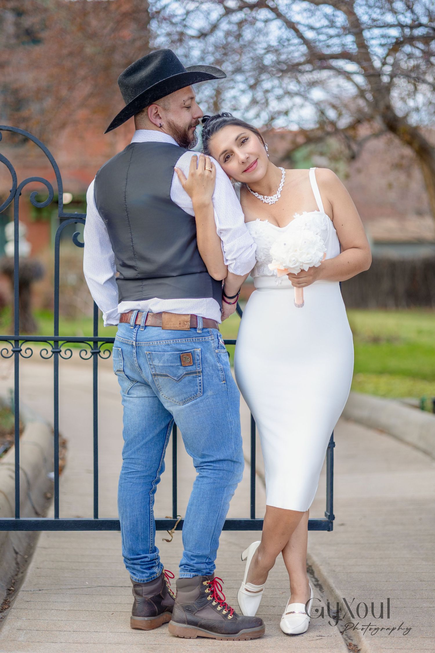 A couple in wedding attire pose together by an iron gate, with her wearing white and him in a vest and cowboy hat.