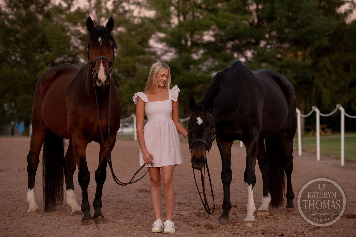 Person in white dress poses with two elegant horses against pine trees at dusk.