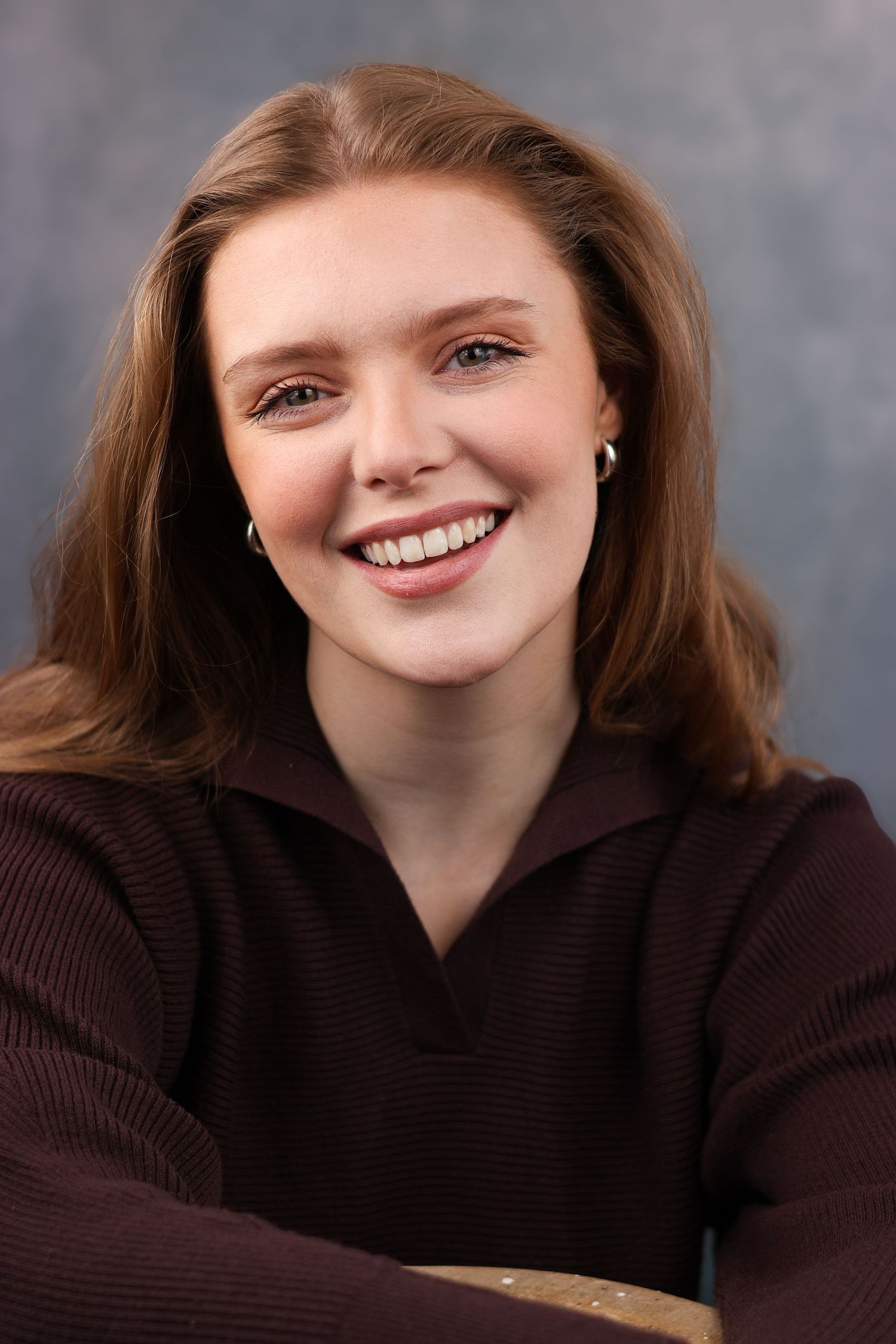 Natural portrait with warm smile wearing brown sweater against grey backdrop.