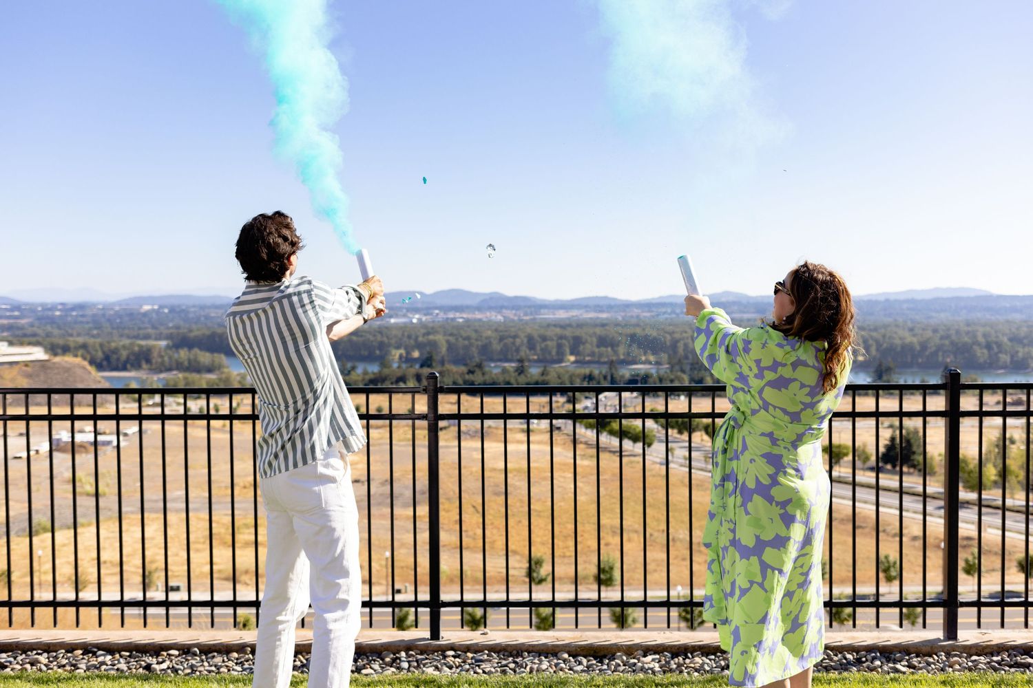 Two people hold colorful smoke flares on a scenic overlook with golden fields and mountains in the background.