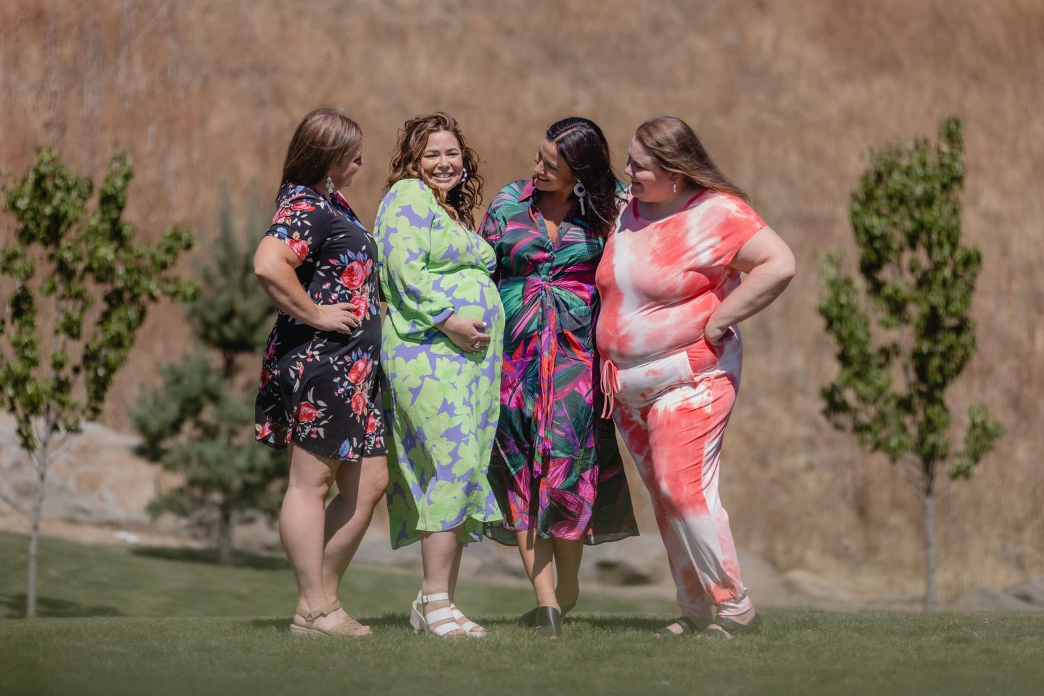 Four friends in colorful floral dresses stand together on grass, chatting and smiling on a sunny day.