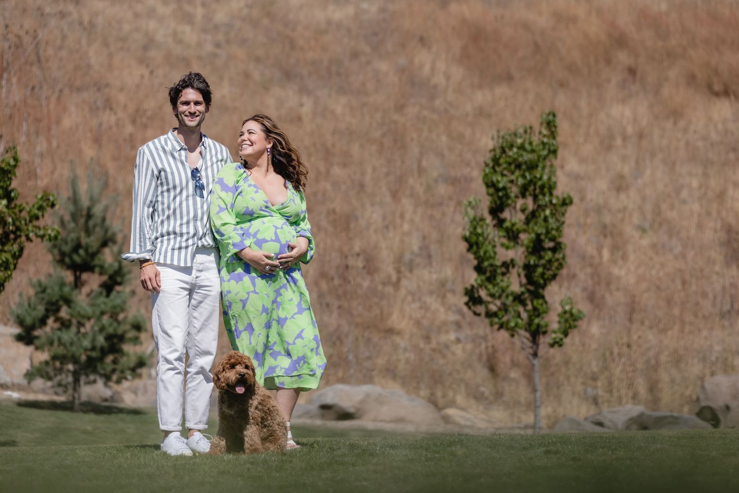 A couple in spring attire stands on grassy lawn with their small dog as young trees grow in the background.