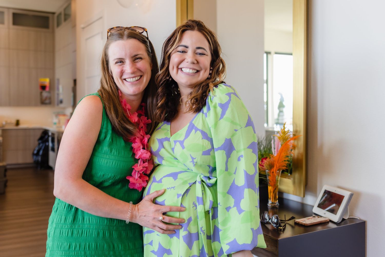 Two women share a joyful embrace while wearing bright green and patterned dresses in a home setting.