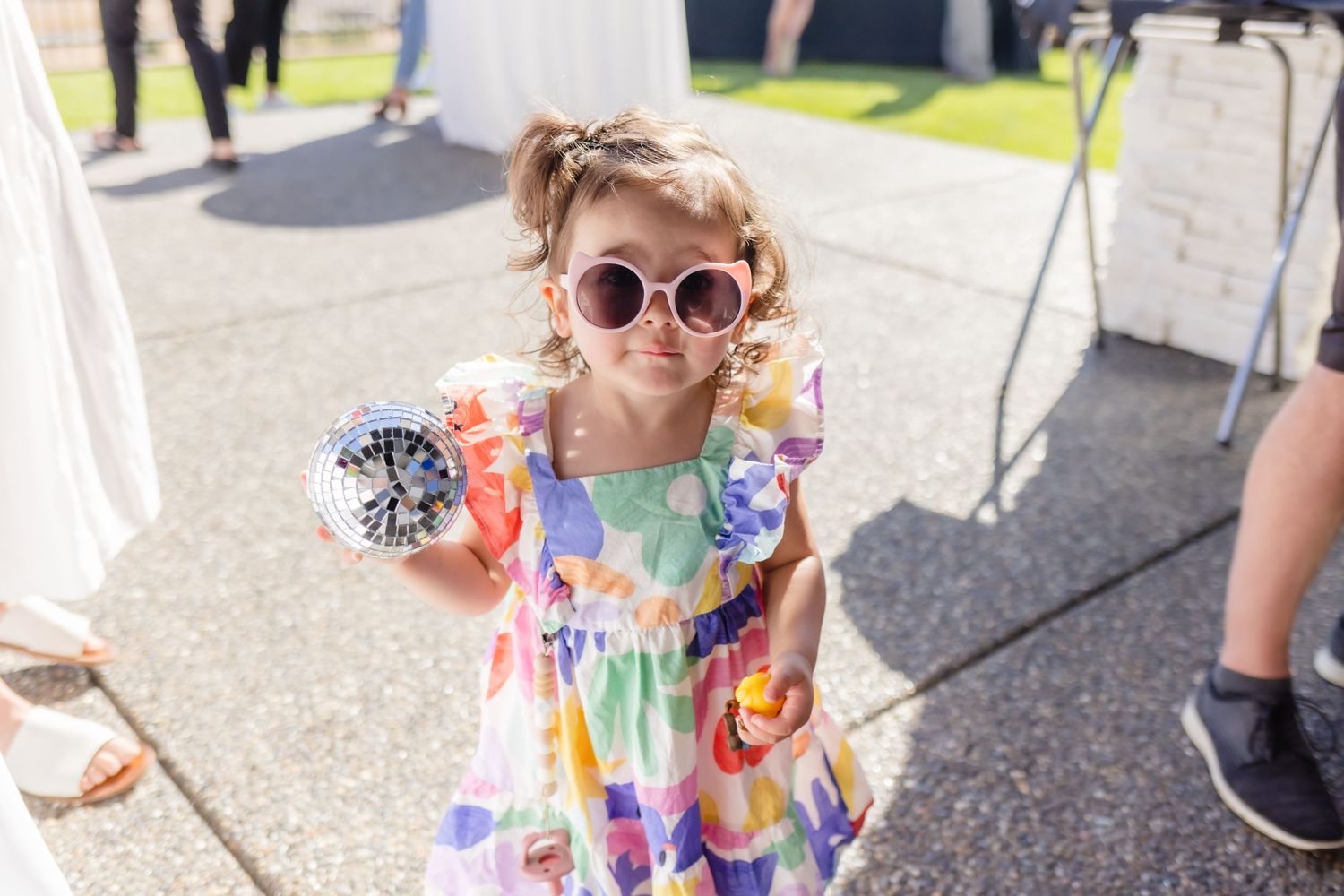 A young child wearing a colorful tie-dye dress and round sunglasses poses on a sunny sidewalk.
