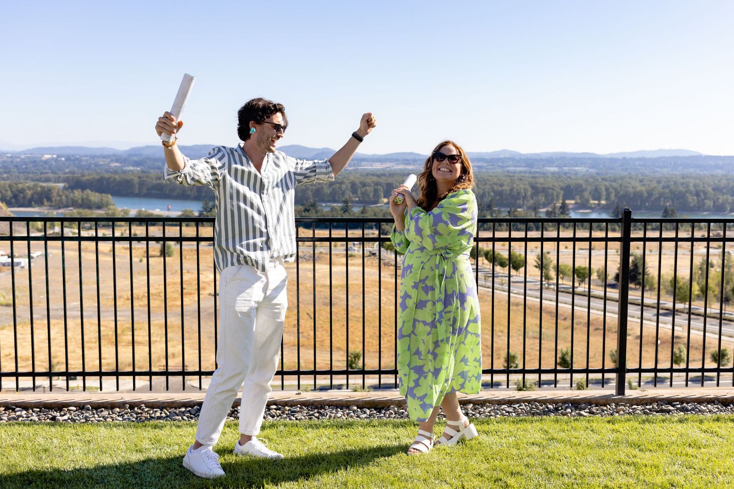 Two people in casual outfits celebrate with raised arms on a sunny balcony overlooking mountainous landscape.