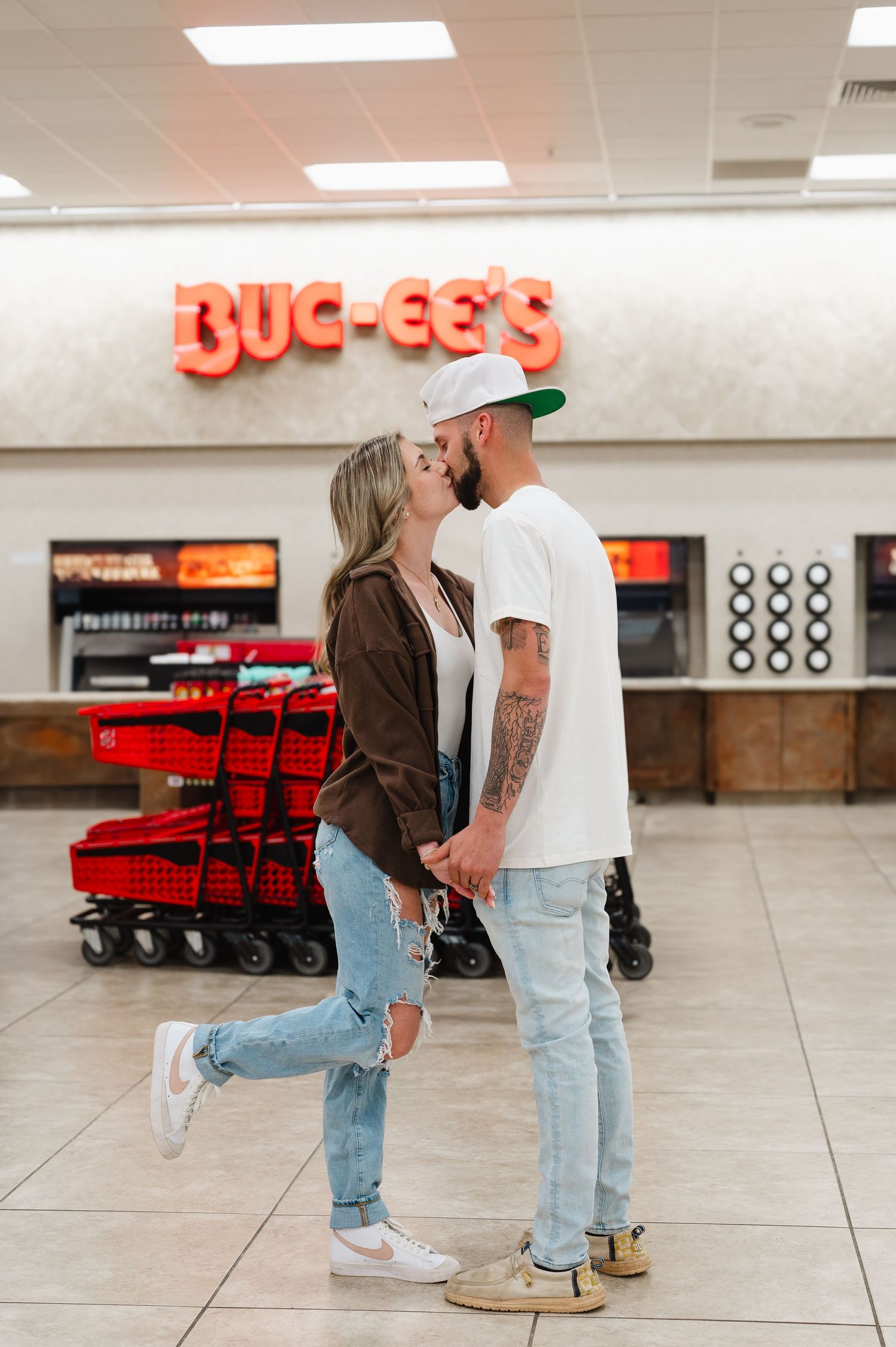 A romantic couple embraces inside a Buc-ee's convenience store with red shopping carts in the background.