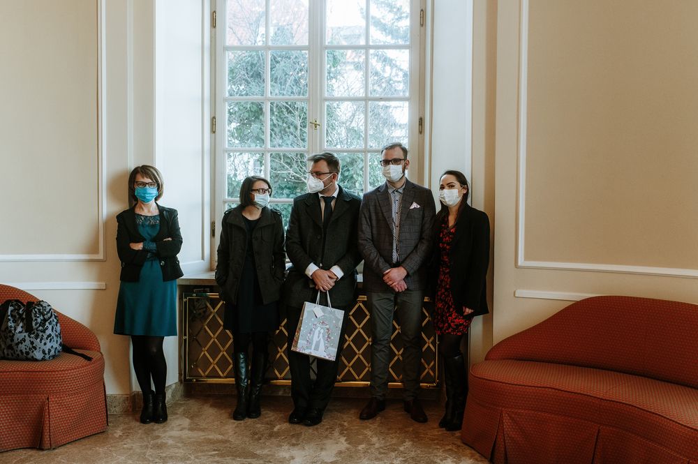 Small group of masked wedding attendees standing against a wall near large windows in formal attire.