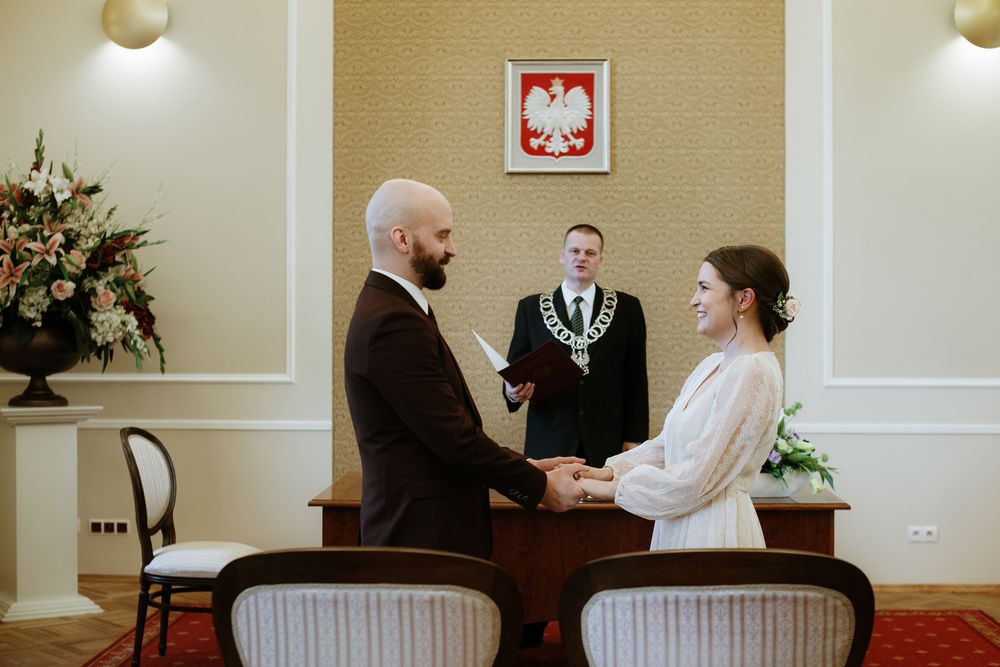 A civil ceremony takes place in a registry office with a Polish national emblem on the wall.