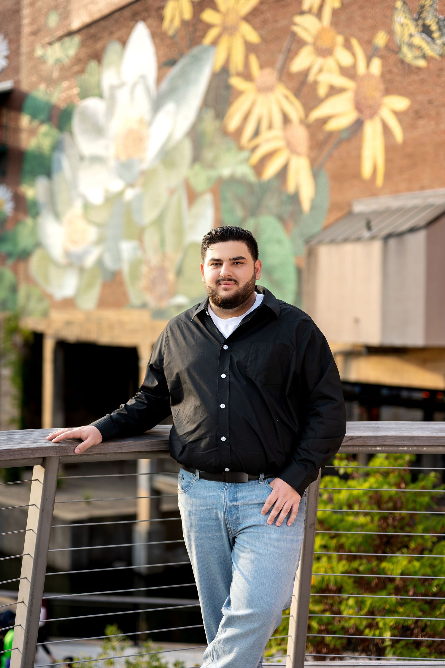 Young man in black shirt and jeans poses on a bridge in front of a colorful floral mural outdoors.