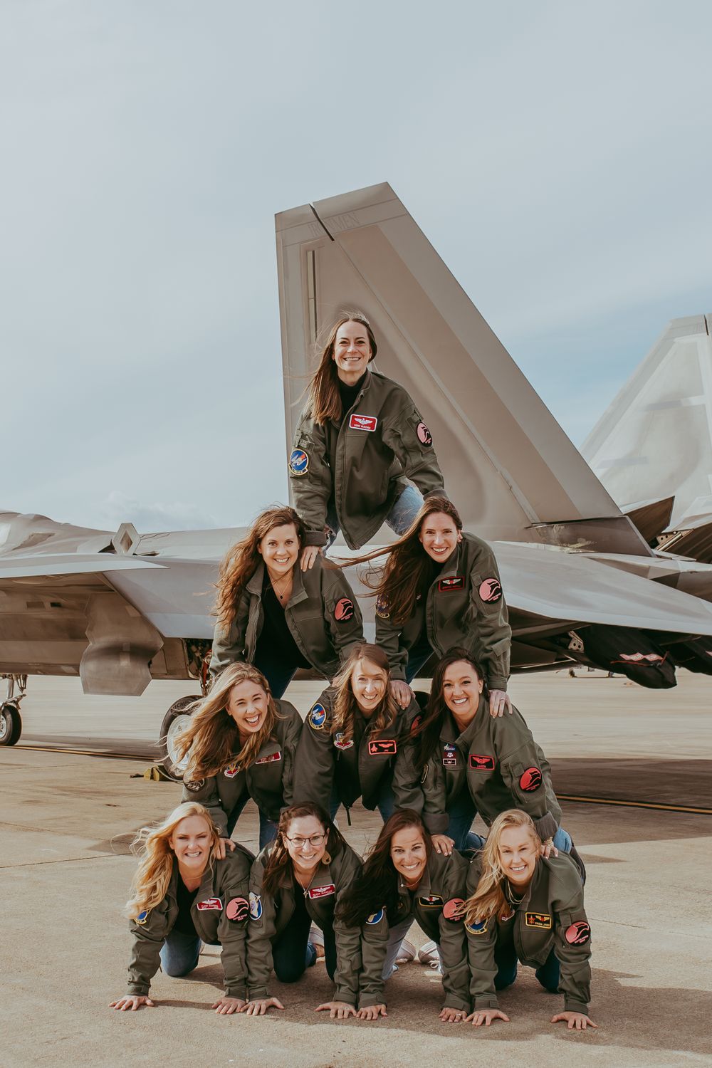 Military aviators in flight suits pose in a pyramid formation in front of a military aircraft.