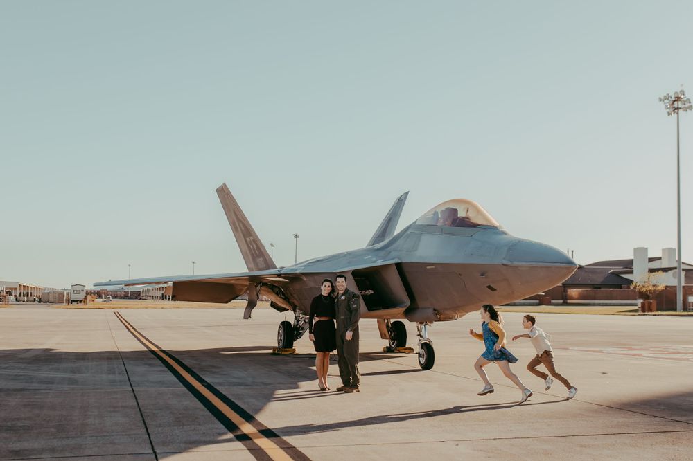 Military personnel and children run alongside a parked F-22 Raptor fighter jet on an airfield tarmac.