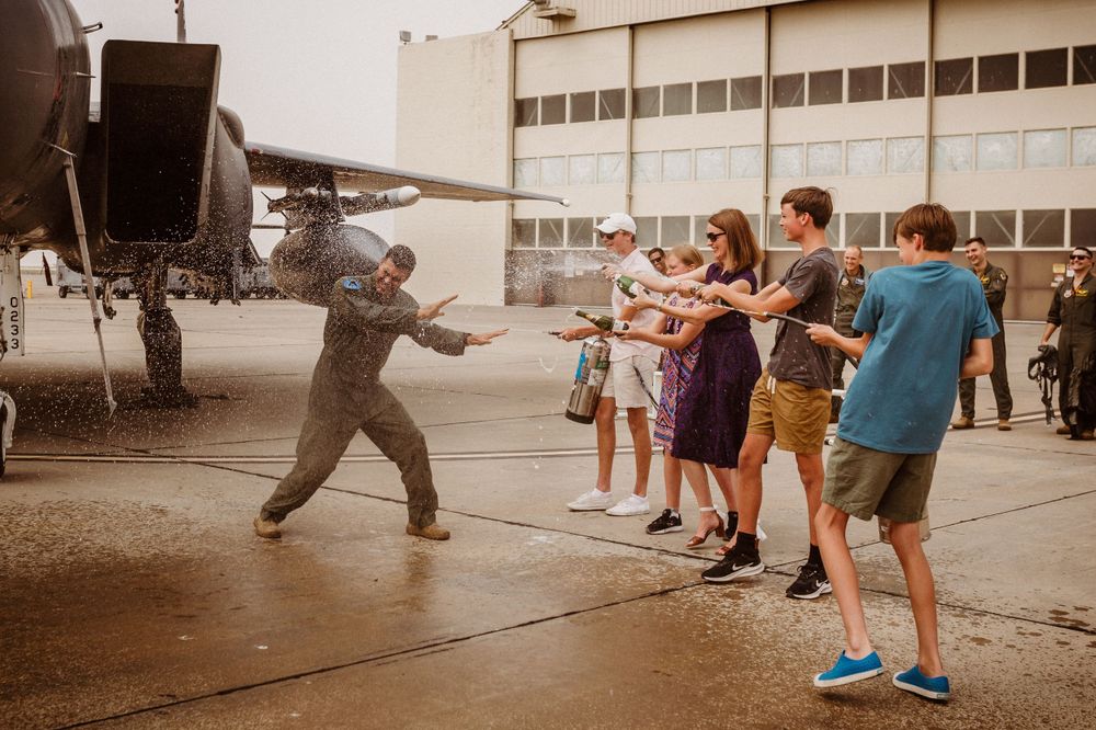 A group of people participate in a fini flight on an outdoor tarmac in front of a hangar.